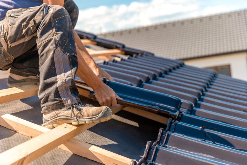 A roofer is installing dark tiles on a new roof under a clear sky. The person, wearing work boots, crouches on wooden beams. The roof is partially tiled with replacement guttering in progress and a house visible in the background.
