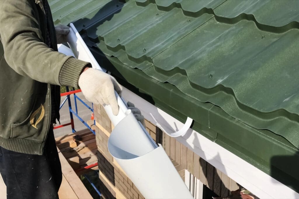 A roofer wearing gloves carefully installs a white rain gutter under a green metal roof on a building. The close-up image highlights the gutter being securely attached along the edge, showcasing expertise in handling both new roofs and emergency roof repairs.