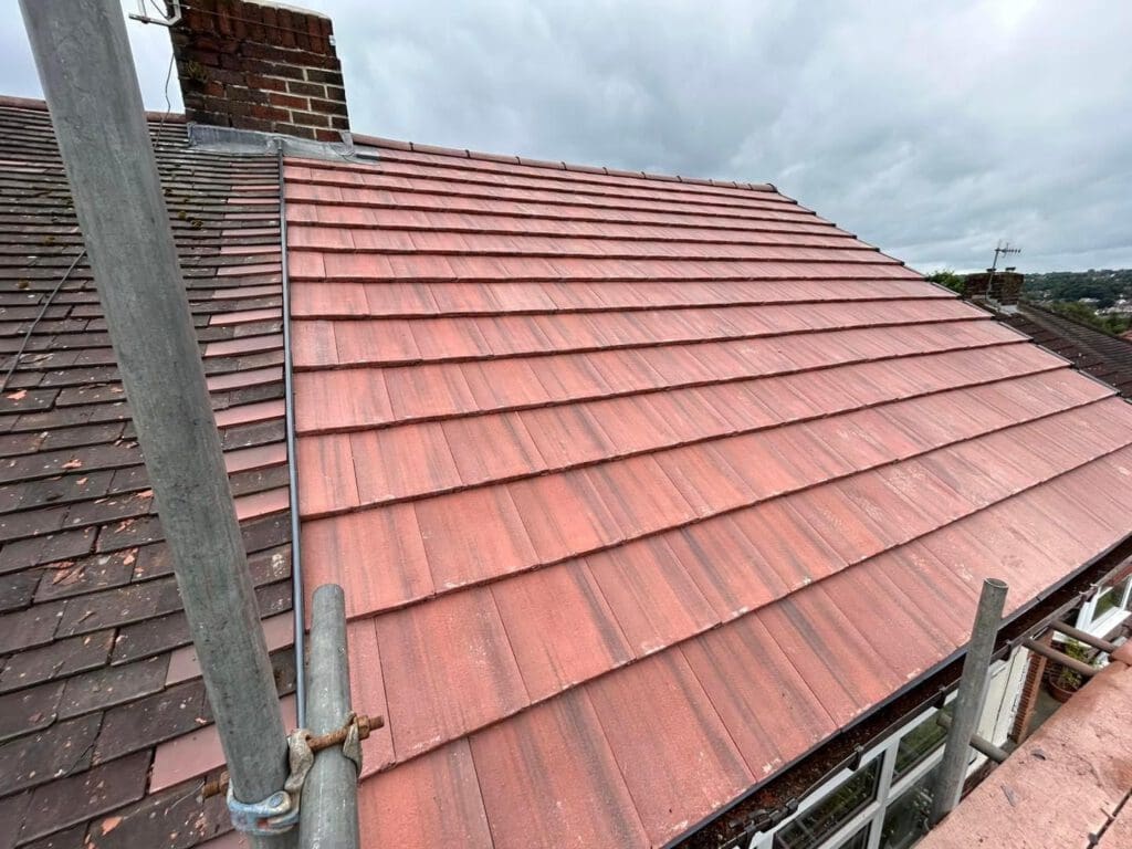 The newly tiled red roof section, crafted by a skilled roofer, sits beside an older brown tiled roof. A metal scaffolding pole rises on the left against an overcast sky, hinting at the possible need for replacement guttering under such moody weather.