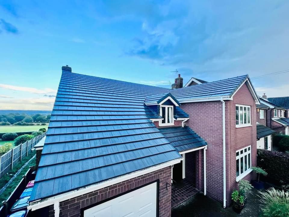 A red brick house with a dark gray tiled roof, expertly crafted by our skilled roofer, featuring a garage and a white-framed window. The sky is partly cloudy, and the background shows green fields and fences.