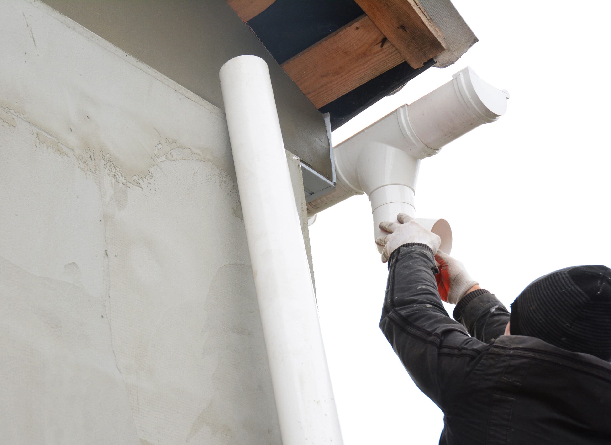 A roofer in a black jacket and hat is expertly installing a piece of white gutter pipe at the corner of the building's roof. Using both hands, they fit the pipe into place on the white wall with its wooden-edged roof.