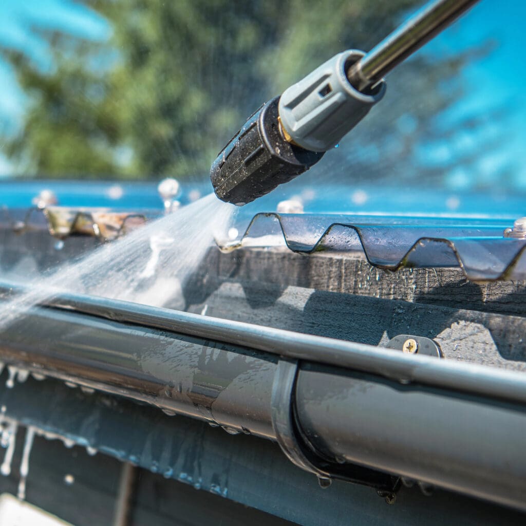 Close-up of a roofer using a pressure washer to clean a roof gutter. The spray of water highlights the cleaning action, with the background out of focus, showcasing greenery and part of the sky—a glimpse into expert care for replacement guttering.