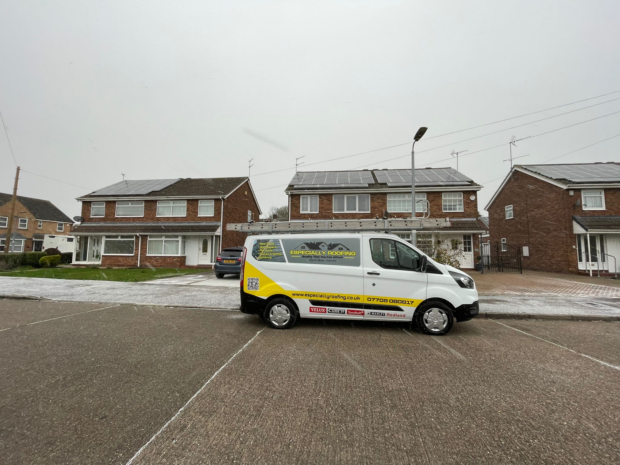 A white van with yellow accents, branded with "roofing," is parked on a residential street. The scene features brick houses adorned with solar panels and overcast skies. The frosty ground sets the stage for discussions on flat roofs and replacement guttering services.