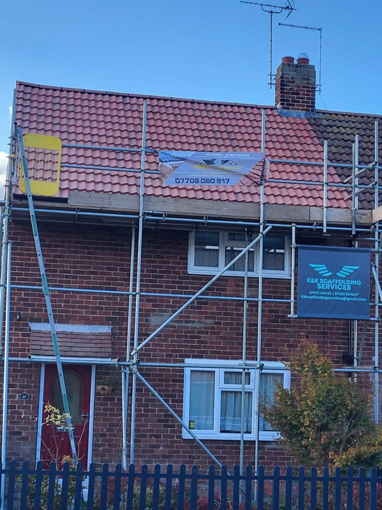 A brick house with scaffolding covering the upper front. Signs for roofing and scaffolding services are attached to the scaffolding. The roof appears recently tiled, and a red door and white windows are visible below.